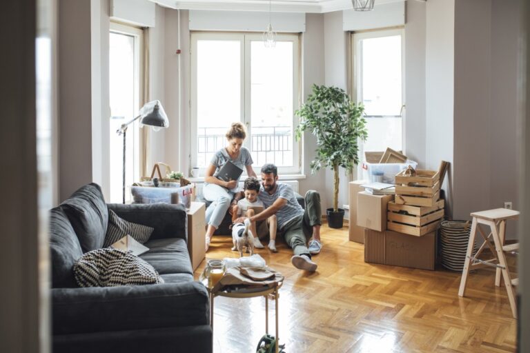 Family sits on a wooden floor with a small dog, surrounded by boxes in a bright room.
