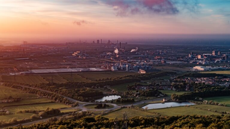Wide view of industrial plant and farmland with buildings, tanks and distant city skyline.