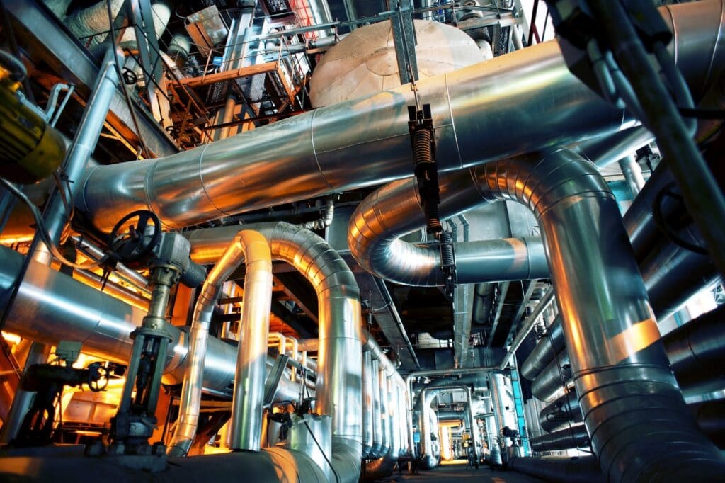 Network of large metal pipes, valves and ducts inside an industrial plant with overhead structures.