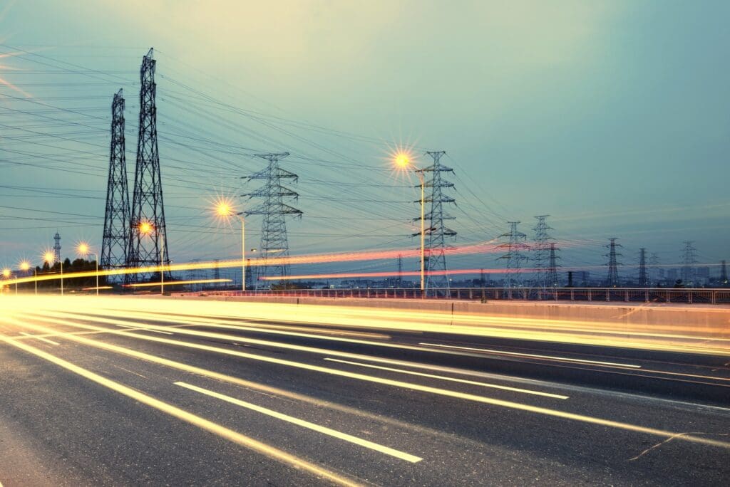 Light trails from traffic pass beneath power lines and pylons along a road at dusk.