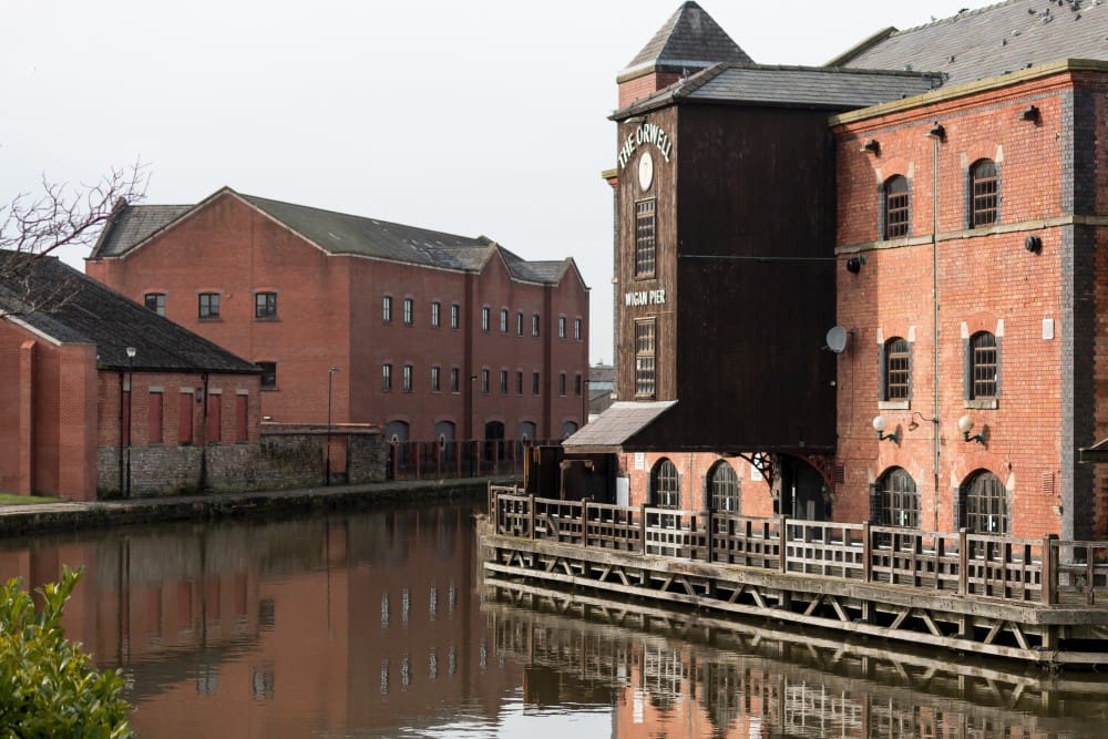 Historic red brick buildings at Wigan Pier reflected in canal water beside a walkway.