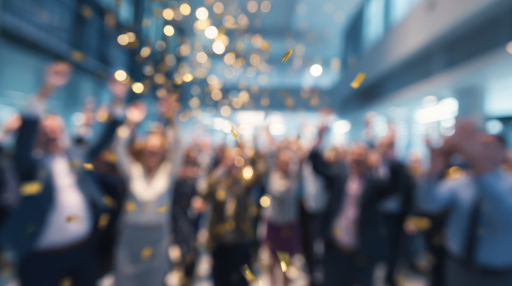 Gold confetti falls over a crowd gathered indoors during a workplace celebration or event.