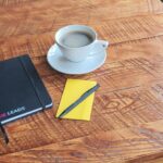 Notebook, pen and coffee cup arranged on a wooden table in a workspace.
