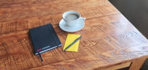 Notebook, pen and coffee cup arranged on a wooden table in a workspace.