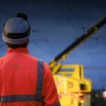 Worker in high-visibility clothing and hard hat beside a yellow rail maintenance vehicle.